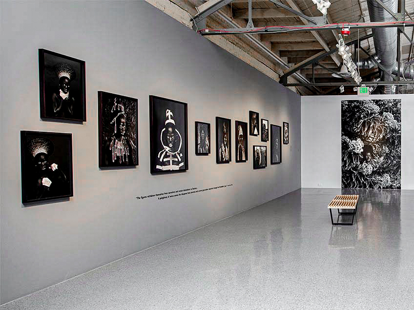 Art gallery with black-and-white photographs on a grey wall, a wooden bench, and an industrial ceiling.