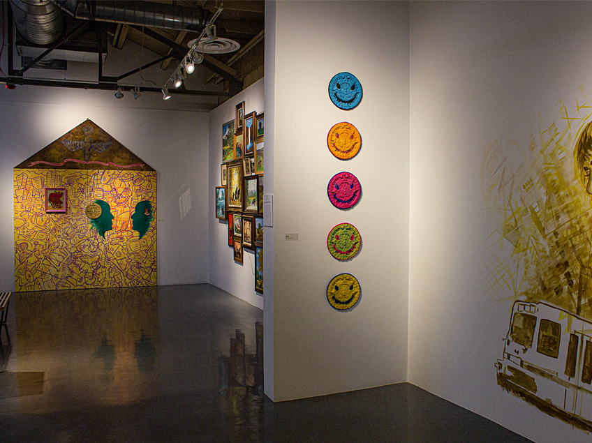 Interior of an art gallery with diverse colorful artworks on display, including a mural, framed paintings, and smiley face art.