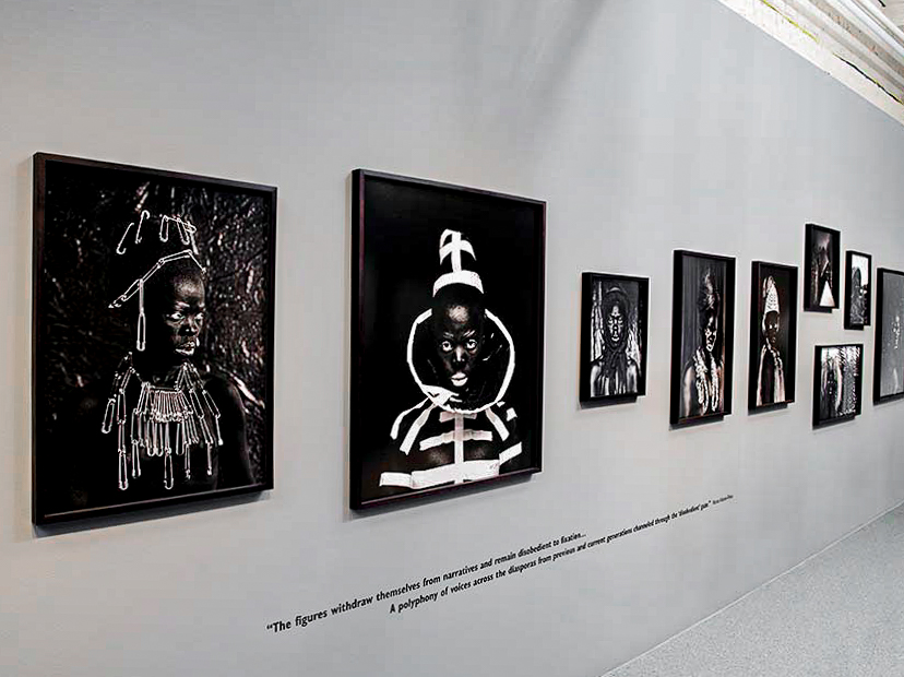 Art exhibit wall with black and white portraits featuring individuals with elaborate headpieces and accessories.