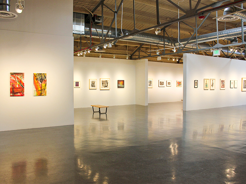 A modern, well-lit art gallery with framed paintings and screen printed images on white walls and a wooden bench in the center.