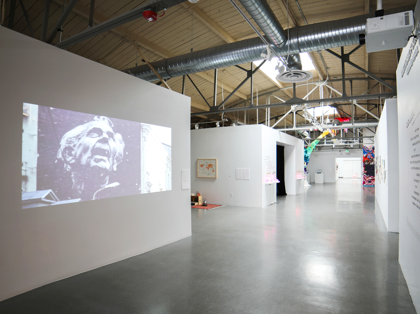 Interior of an art gallery with an industrial design featuring a projected image of an older man’s face and various artworks.