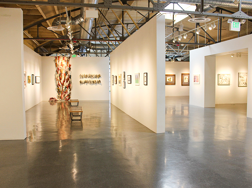 A modern art gallery with white walls, framed artworks, a floor-to-ceiling sculpture, and benches on a polished concrete floor.