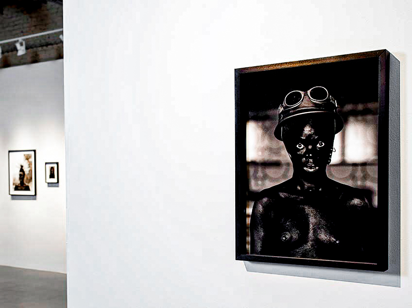 A black and white portrait photograph in a gallery, showing a person with goggles and bare torso.