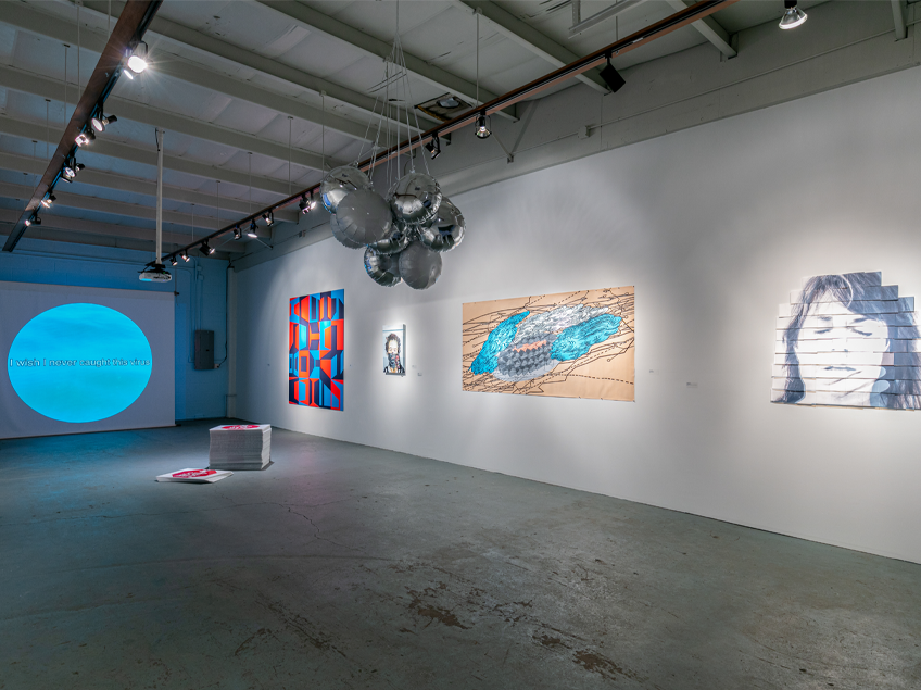 Gallery view with a video projection on the wall, accompanied by a brightly colored graphic painting in red, blue, and purple. Nearby is a portrait featuring a face mask, along with an abstract painting in blues and tans with line work, and another face painting. On the floor is a pop art installation.