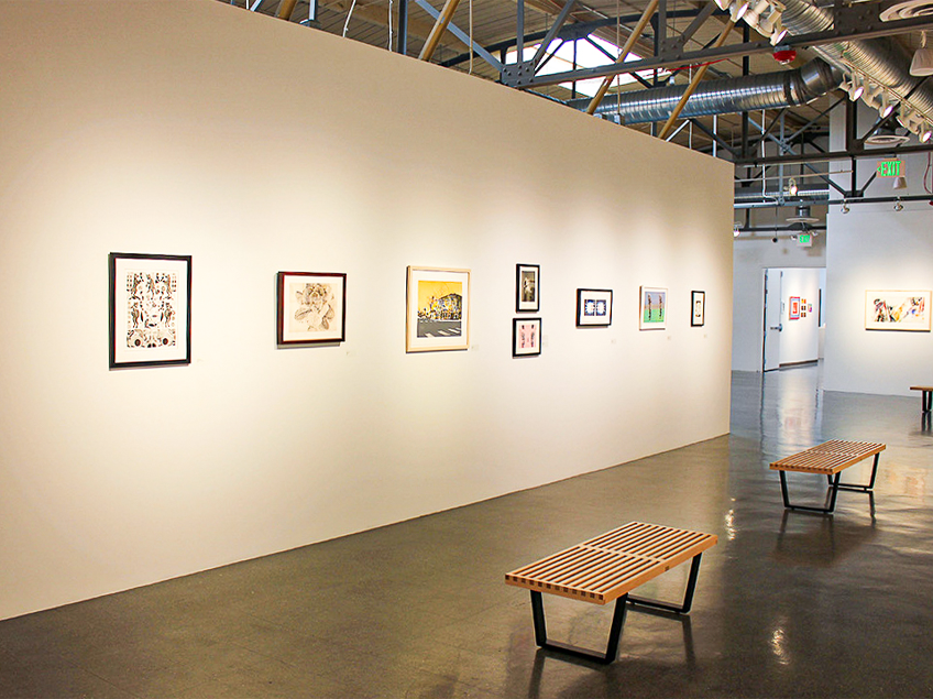 Art gallery interior with white walls displaying framed artworks and two wooden benches on a concrete floor.