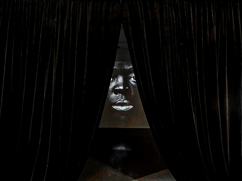 Dark curtains parted to reveal a projected face within the room.