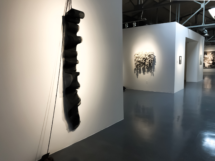 Art gallery interior with black vertical sculpture and textured white artwork on walls.