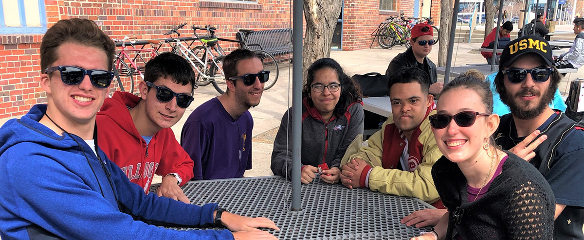 IHES students seated around an outdoor table, most wearing sunglasses and smiling for the camera.