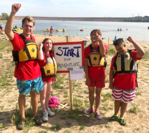 IHES students at a beach at a lake wearing floatation vests and standing with fists in the air around a sign that reads "Start Your Adventure".