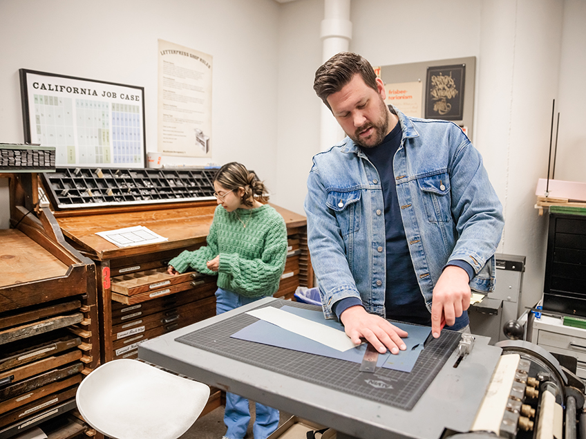 Students work in the Letterpress Studio in Communication Design. You can see the many slender drawers and cubbies which hold the individual letters to use in the Press.