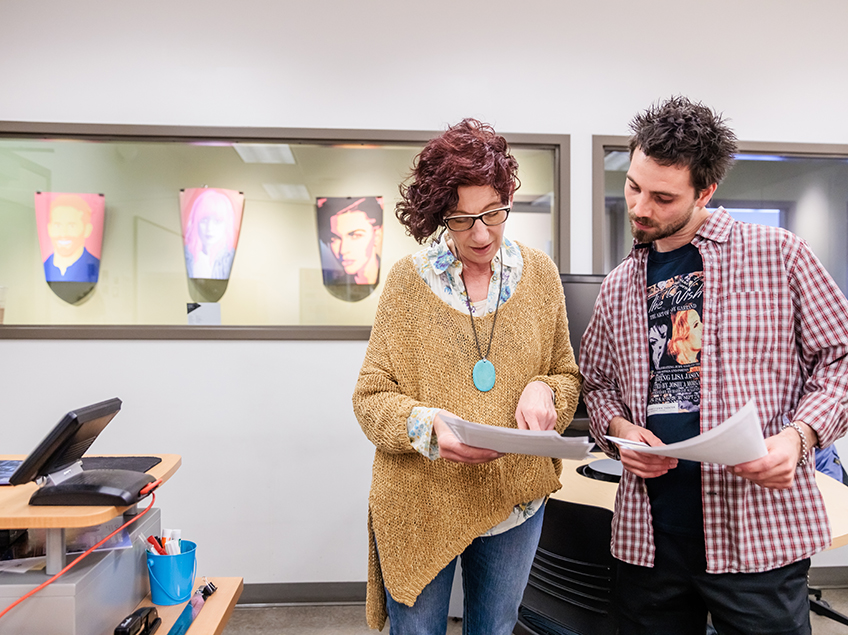 Professor Heidi Cies consults with a student about their final project. Posters on display in the background show graphics of larger-than-life portraits the students created.