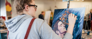 Looking over the shoulder of a student painting on an easel.