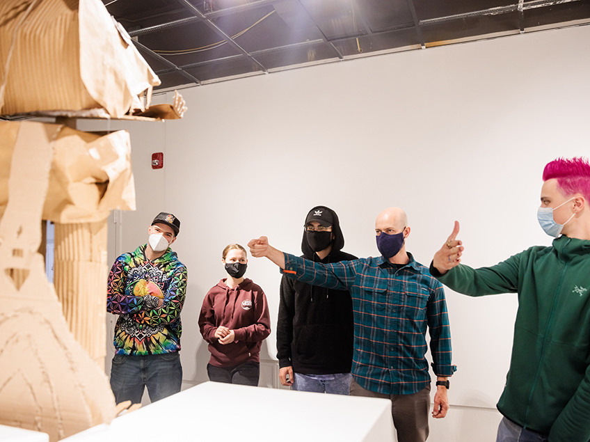 Professor Michael Bernhardt walks through a critique with his class in Gallery 199. By their hand gestures, they are discussing certain angles of the cardboard sculpture on a pedestal in the foreground. One student has electric pink hair.