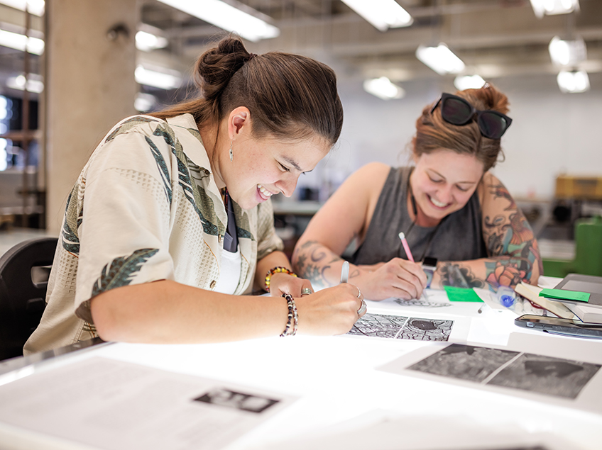 Two students smiling and laughing while working on the light table in the Print Studio.