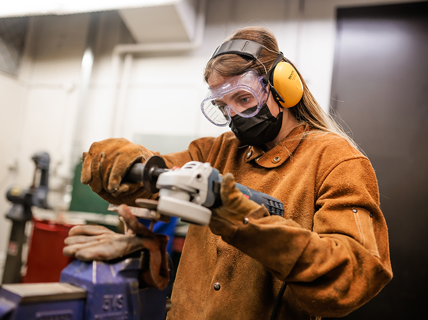 A student works with heavy machinery while wearing a heavy leather protective smock and gloves, along with soundproof headphones and safety goggles.
