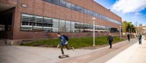 Brick building is the exterior of the Art Building, and a student is skateboarding by.