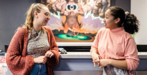 Two women talk to each other in front of a desk.