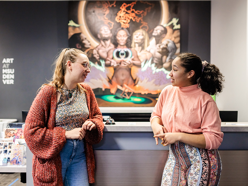 Two students chat while standing at the front desk of the Art Deparment. Behind them is a painting of a diverse group of individuals representing the many peoples of earth. They hold a central floating orb and cube, in poses of reflection and contemplation.