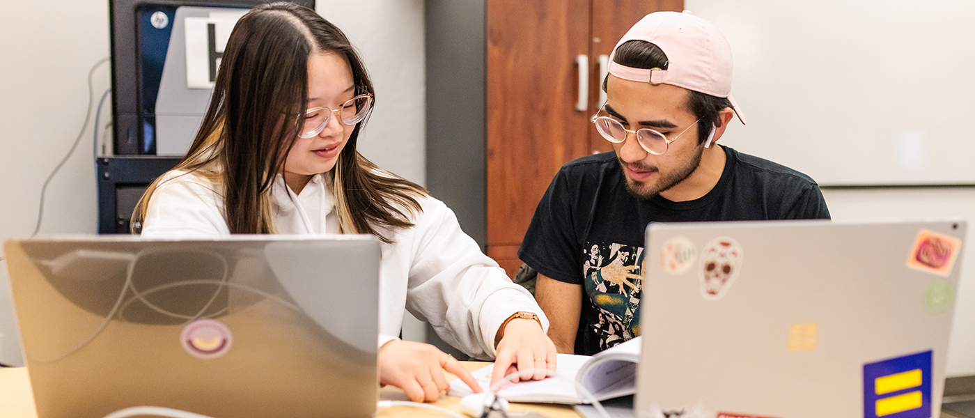 Two students sitting next to eachother to collaborate, each with thier own MacBook Pro laptop to work on.