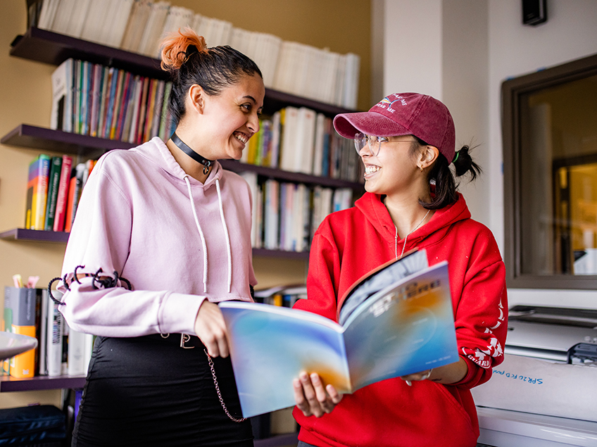 Two Communication Design students flip through the Metrosphere magazine, which they helped to design and create. They stand in the CDES library in front of the many books and periodicals which are available for student research and reading.