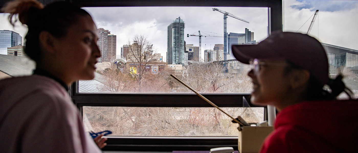 Two students talking in CDES Suite, with view of Denver skyline seen through a window in the background