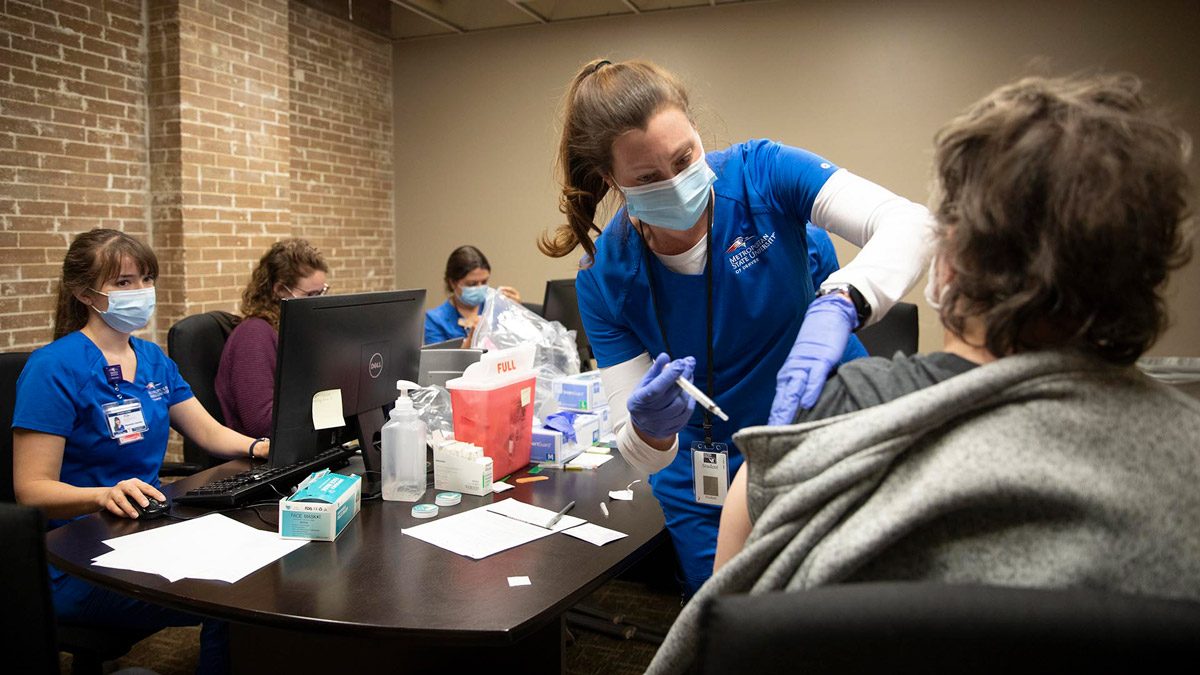 A nursing student giving a shot to a patient in a room with a table and several other individuals watching