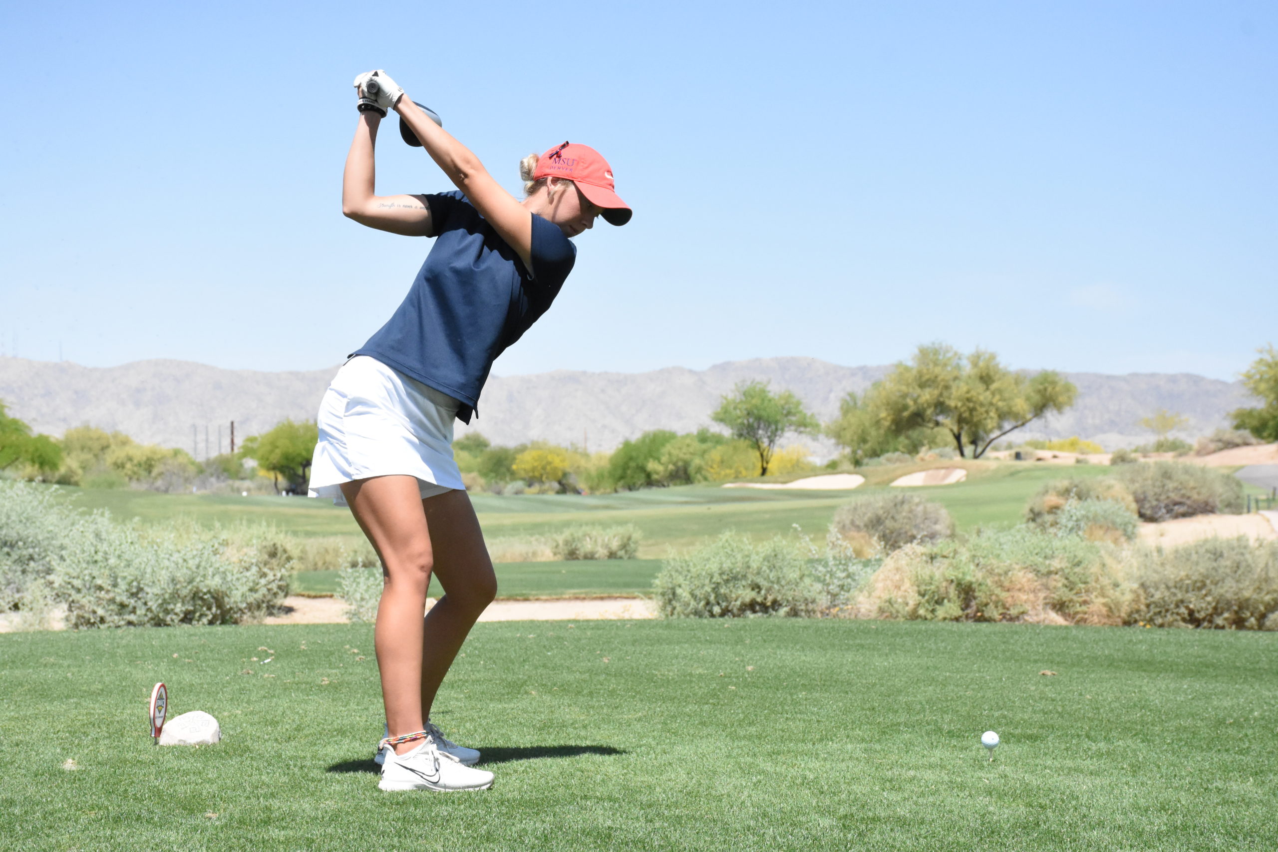 Golf athlete Courtney Lawler tees off at a desert golf course with a mountain range and trees in front of her.