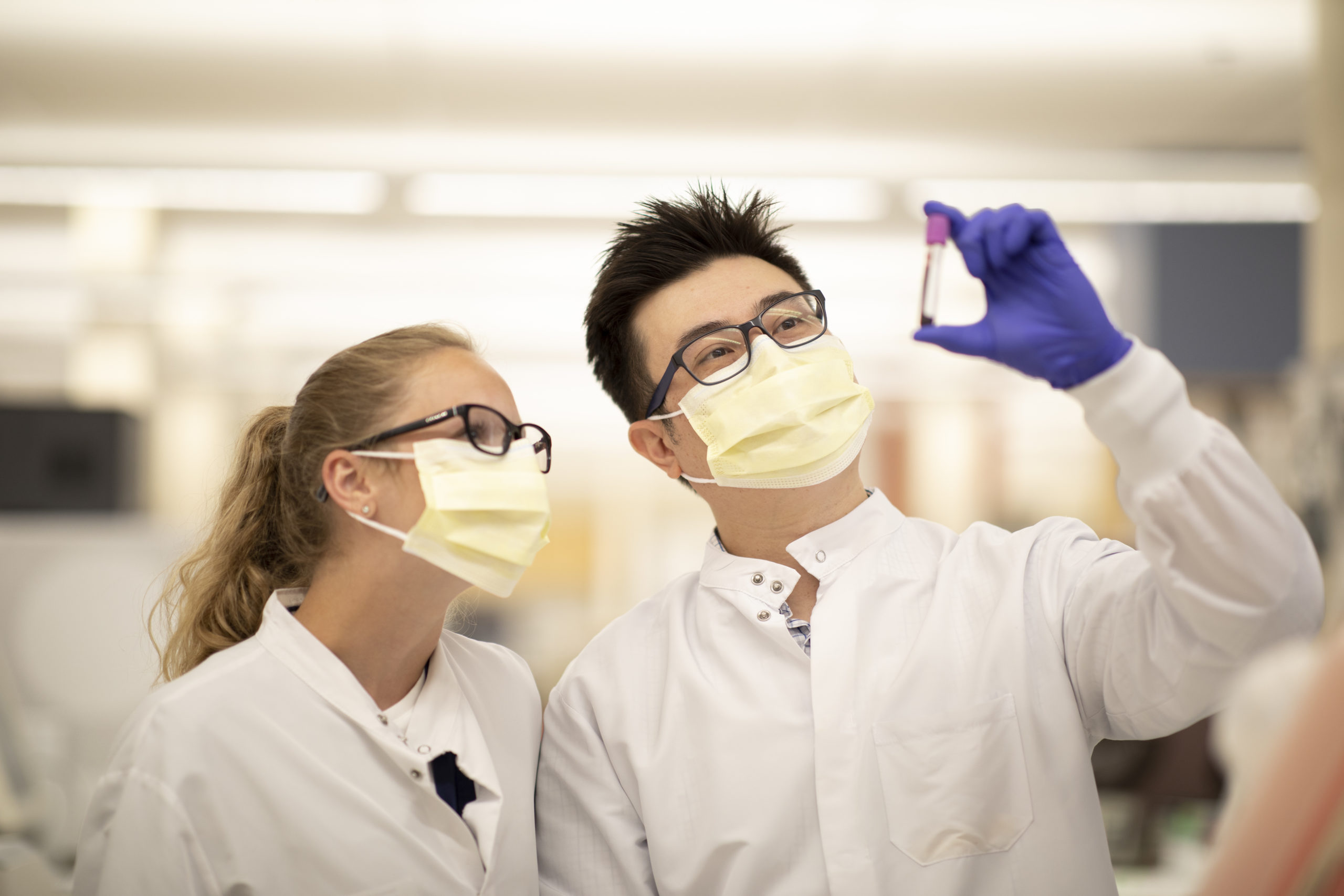 Two people in lab coats and masks examining a test tube sample.