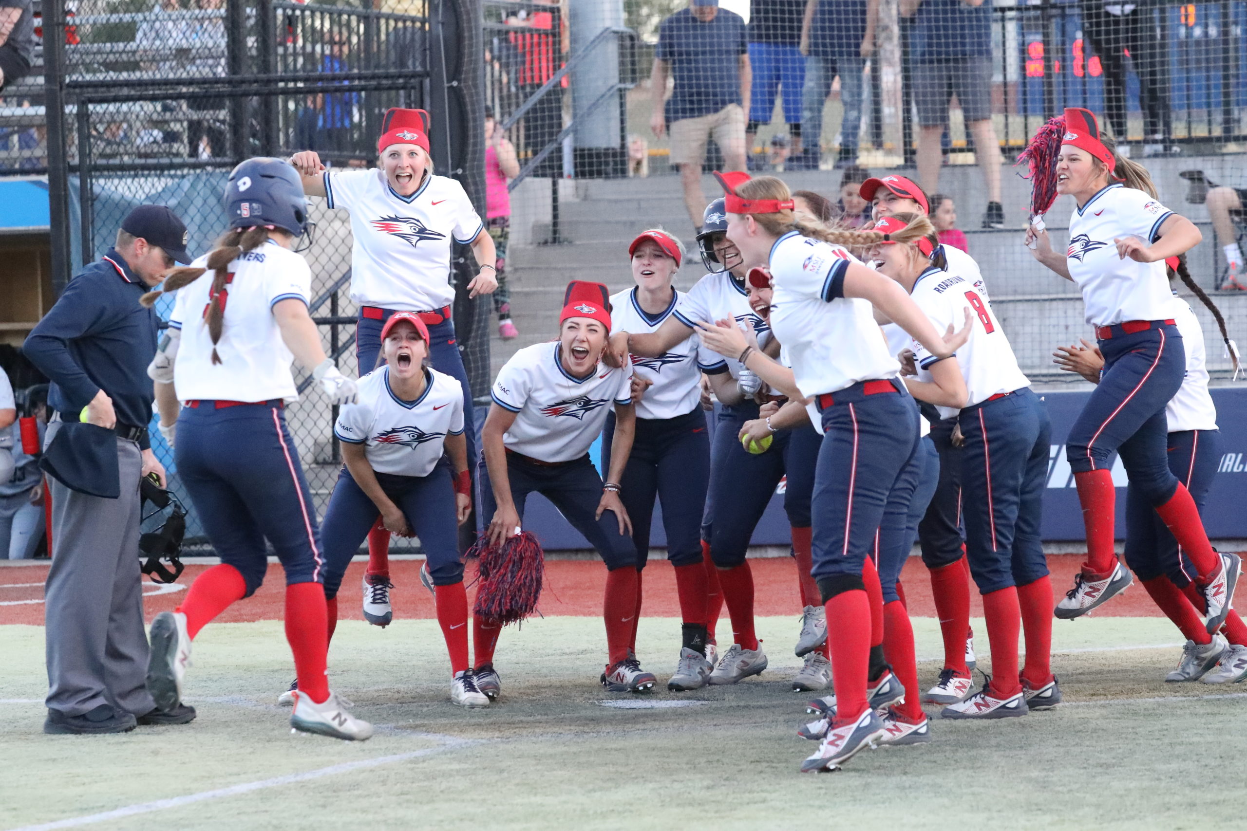 Softball player Shelby Robb approaches home plate after a walk-off home run as her teammates enthusiastically wait to greet her.