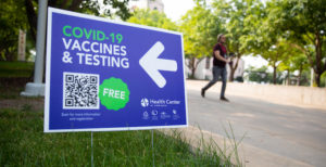 Blue yard sign in grass that says Covid-19 vaccines and testing with a left arrow.