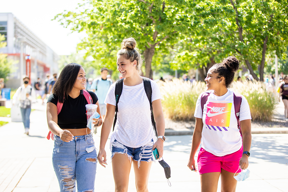 Three students walking towards the viewer, through a green and sunny Auraria campus