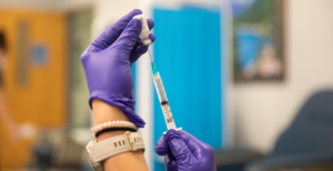 Person wearing purple gloves extracts a vaccine from a vial with a syringe.