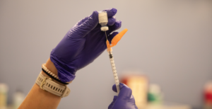 Purple glove (person not pictured) extracts a vaccine from the vial to the syringe.