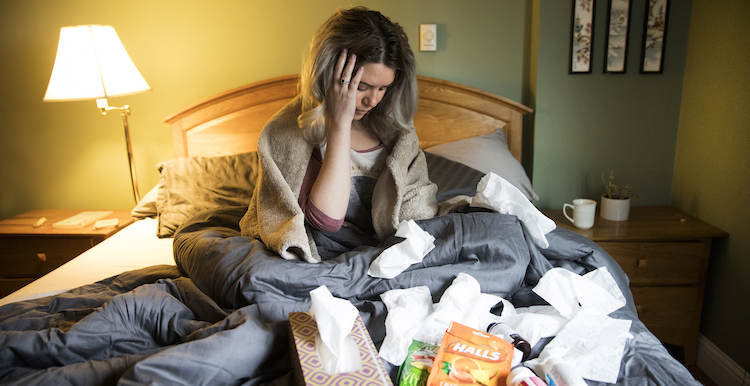 Person sits on bed with tissues, cough drops, medication and more.