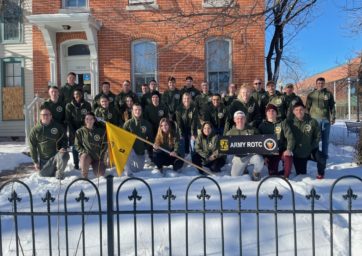 ROTC students posing for a photo of a building on 9th Street Park with an Army flag
