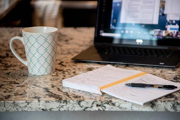 A laptop, notebook and a cup of coffee on a kitchen counter.