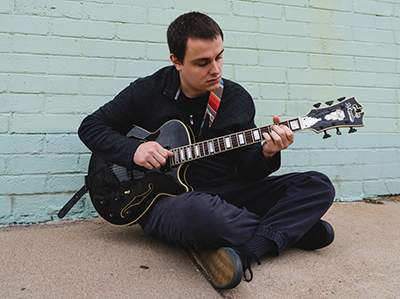 Eric Welty Rodriguez sitting on sidewalk playing guitar