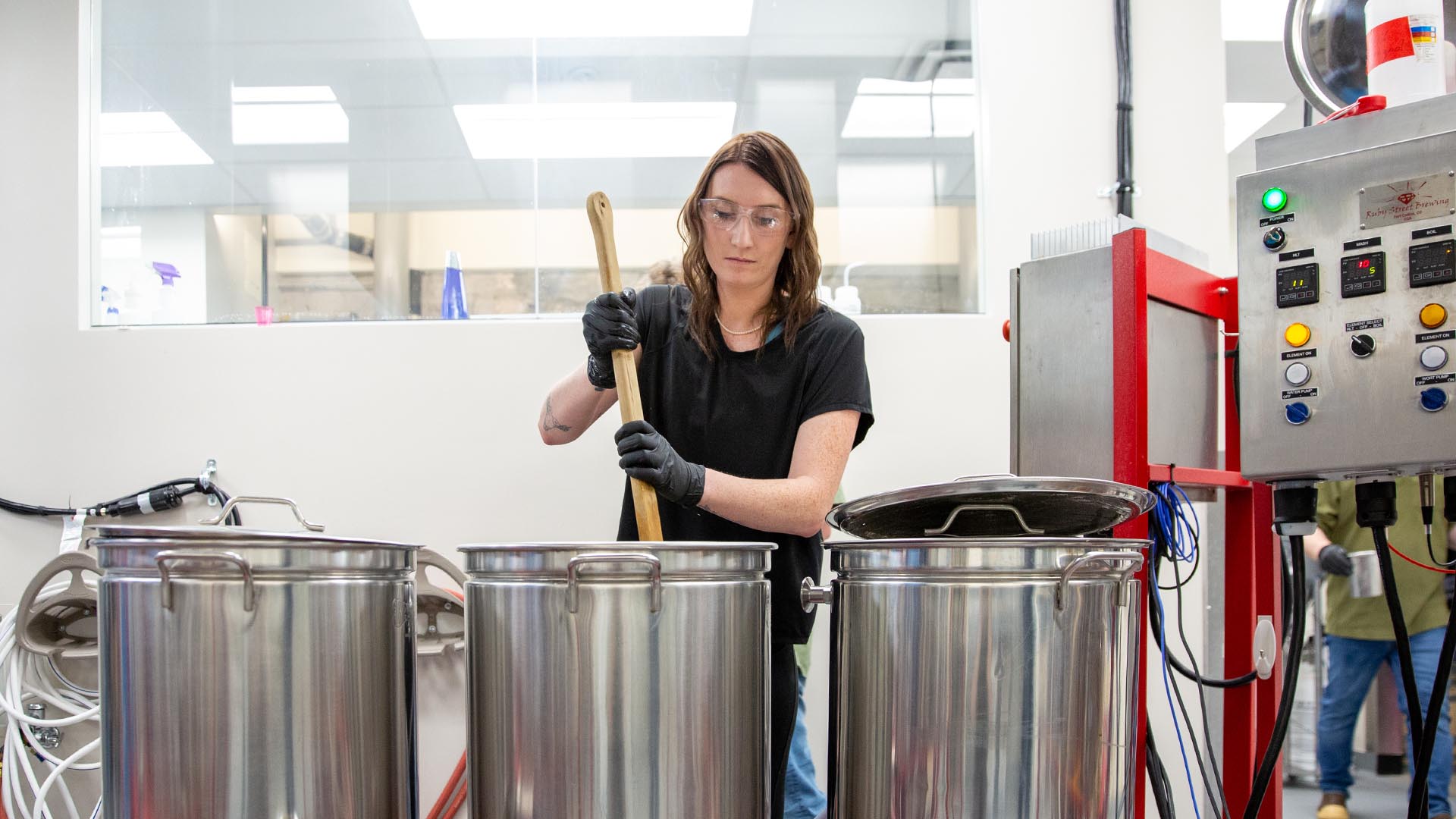 Female student stirring a large, metal pot in a brewery.