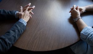 Zoomed in shot of two sets of folded hands on a wooden table.