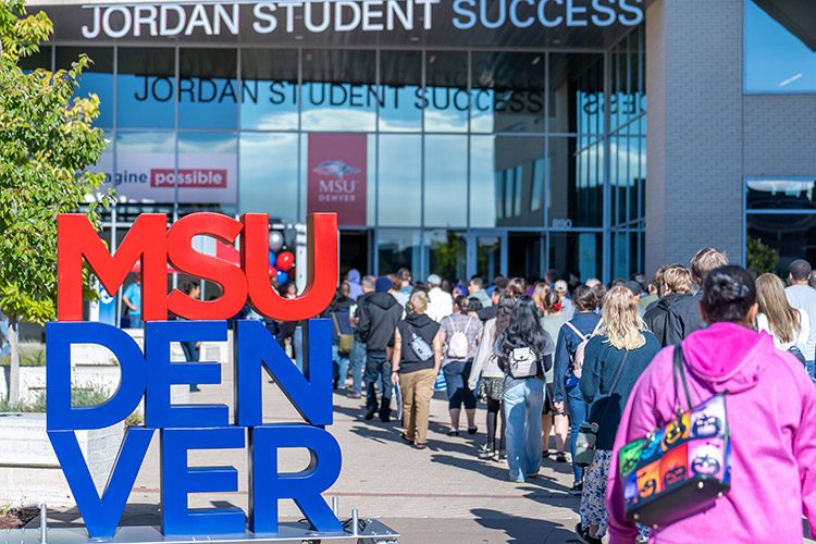 A large number of students walking into the JSSB building with the red and blue MSU Denver sign placed on the sidewalk.