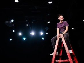 Student Caden Pazo sits atop a ladder, smiling, with theater lights in the background