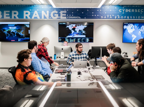 Students sitting around table in the Cyber Range; flat panel monitors hang on the wall behind them