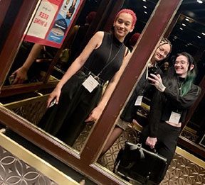 Three students photograph their reflections in a mirror, they are wearing conference badges.