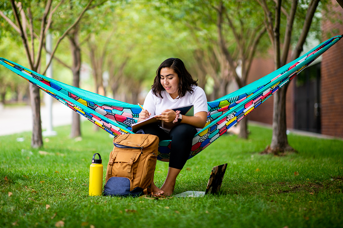 Student sitting in hammock between trees on Auraria campus, with notebook in lap and laptop water bottle and backpack on ground next to them