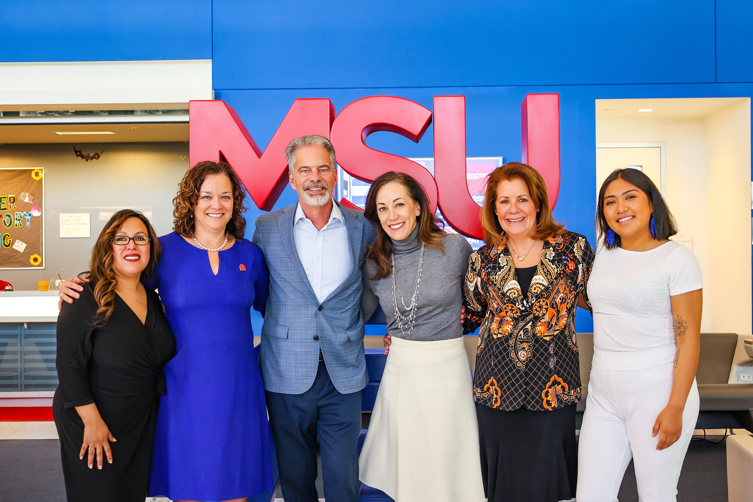 (L-R) Cynthia Baron, Christine Márquez-Hudson, Ferd Belz, Janine Davidson, Christy Belz, and Mariana Pascual-Miranda, standing in front of the red and blue MSU Denver sign in JSSB.