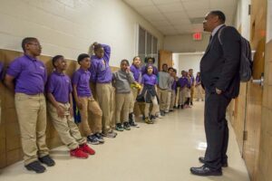 A group of elementary school children are lined up along the wall in a school hallways talking with a Call Me MISTER teacher candidate who stands opposite them.