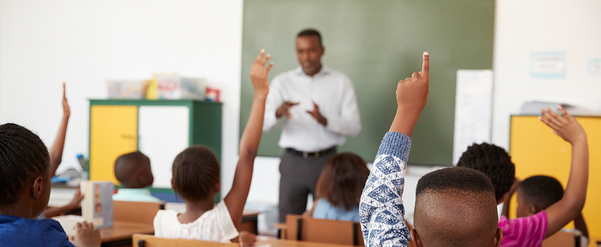 Image of Elementary school students raising their hands with a slightly blurred image of an African-American male teacher stands in front of the class.