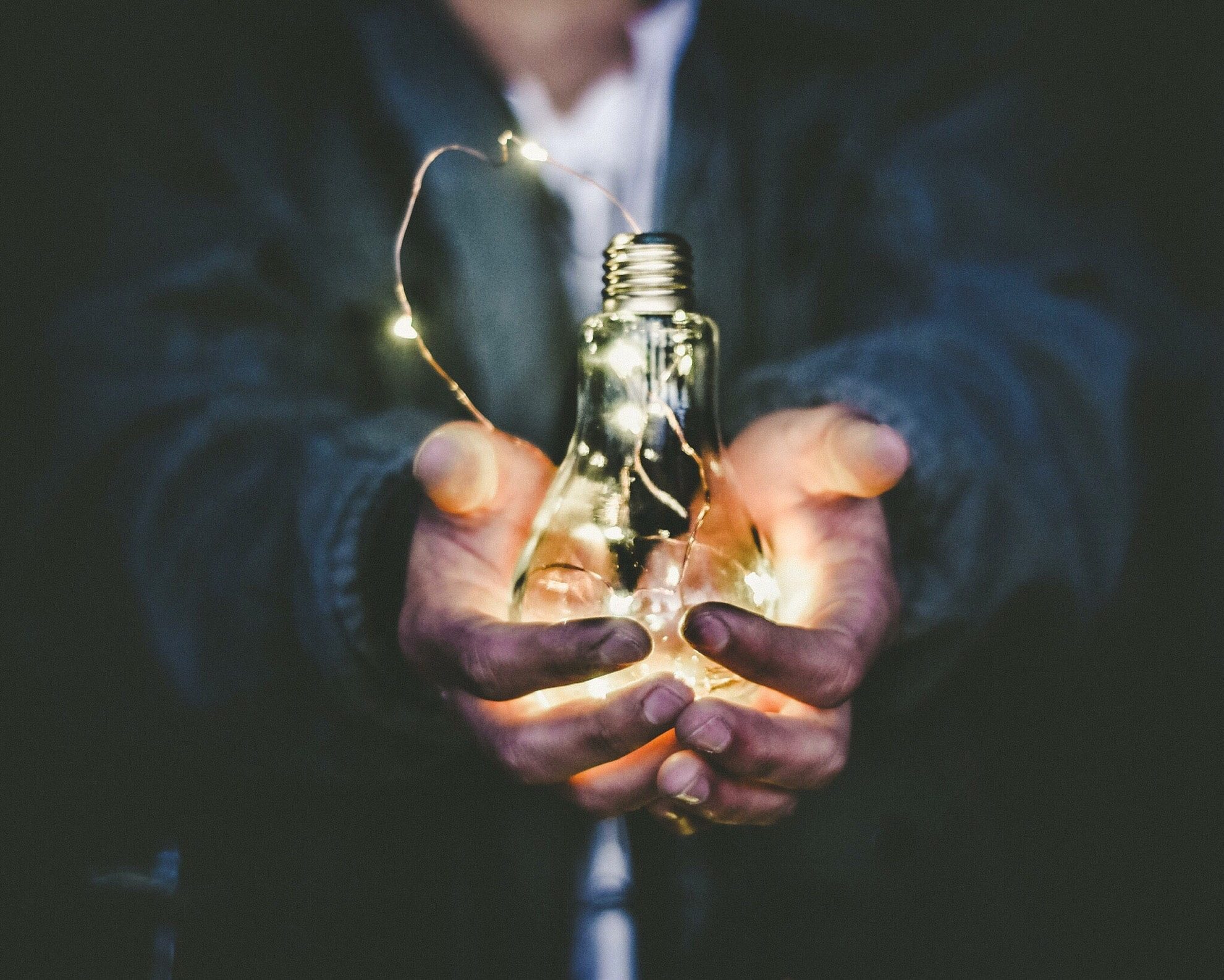 A pair of hands holding a light bulb with a string of lights illuminating the bulb