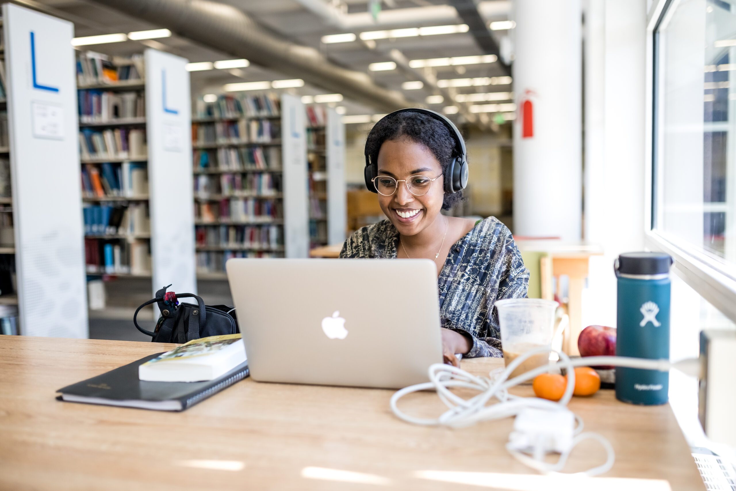 Image of a student sitting in front of a computer with headphones on in the library, smiling.