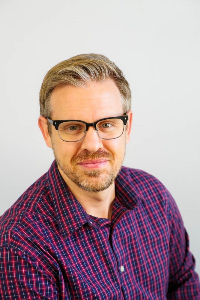 Dr, Gregory Seibert posing facing camera, red checkered shirt with white backdrop.